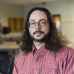 Professor Jonathan Gaffney, with glasses, beard and hair to his shoulders, dressed in a red shirt, stands in his classroom.