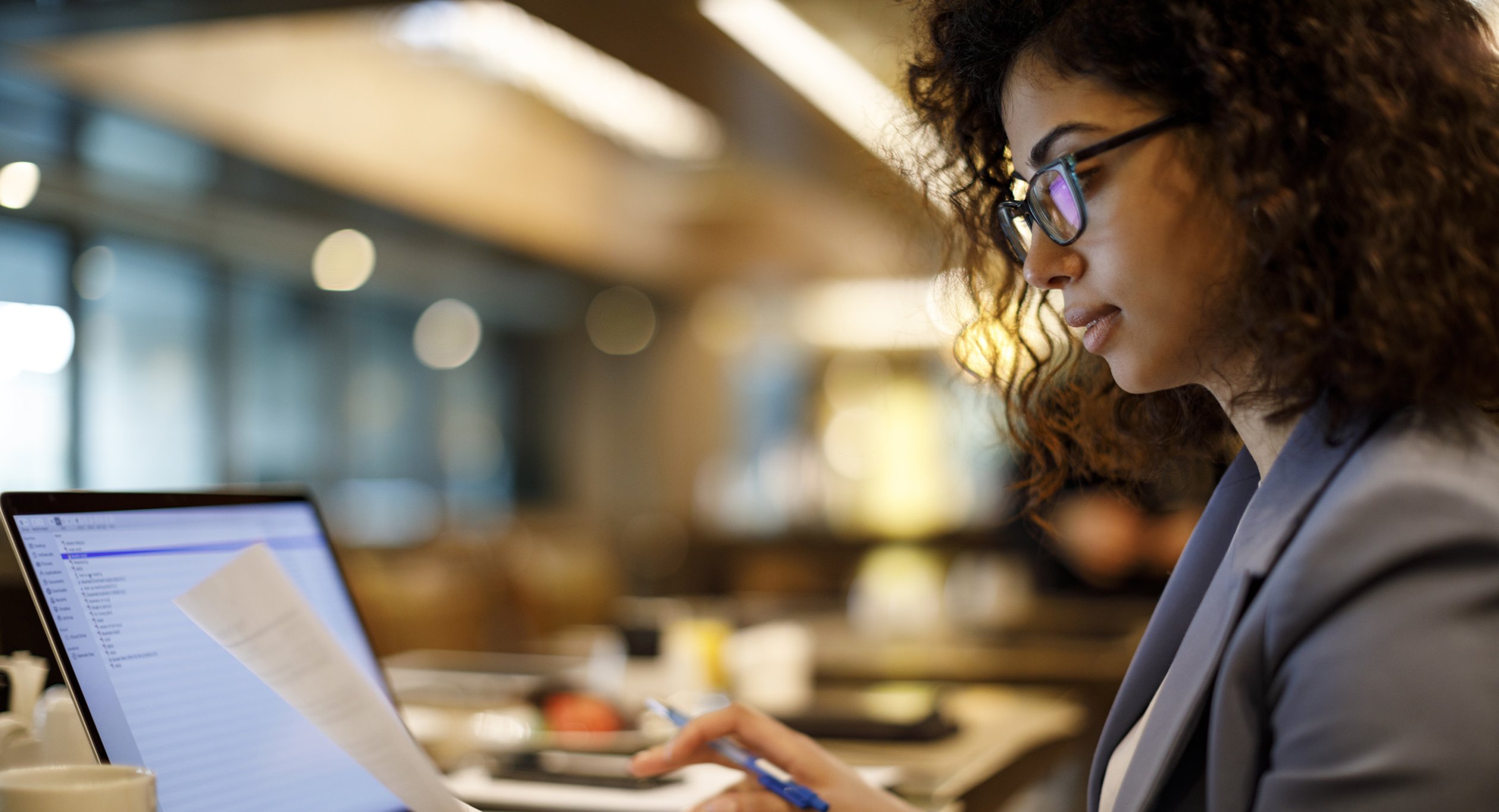 Woman in suit jacket and glasses holds a sheet of paper in one hand while typing on a laptop with the other hand.
