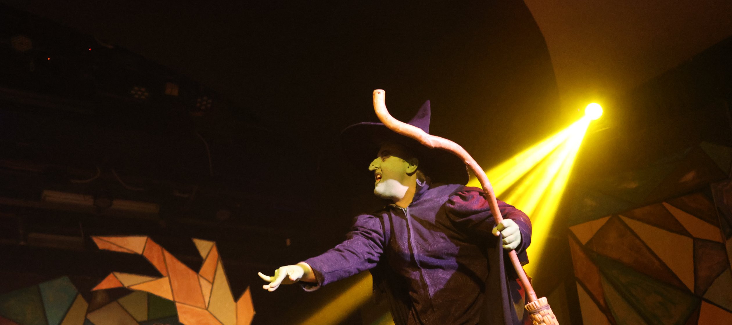 The Wicked Witch of the West holds her broom in one hand and points with the other as a ray of yellow light shines from above.