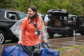 A student wearing a rain poncho carries two large blue bags into a residence hall amid rain on Move-In Day 2025.