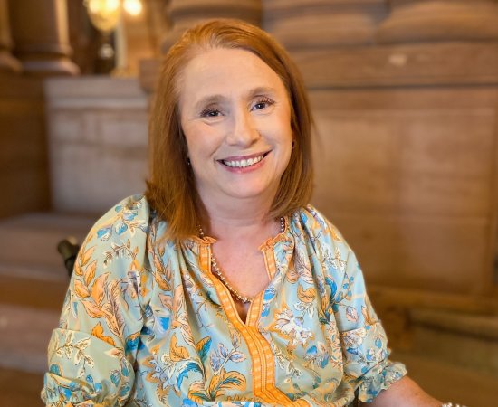 Kimberly Hill, with red hair and a blue-yellow shirt, sits in her wheelchair inside the capital building in Albany, smiling.