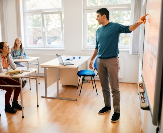 A teacher points to the board at the front of a classroom as students at desks listen.