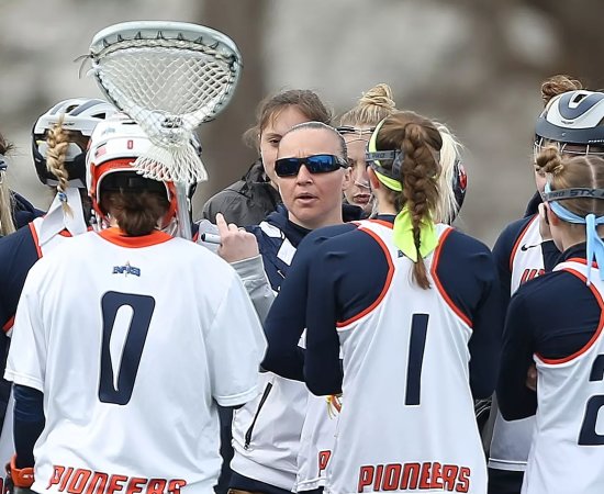 Kristin St Hilaire wears sunglasses and talks with a group lacrosse players on the field.