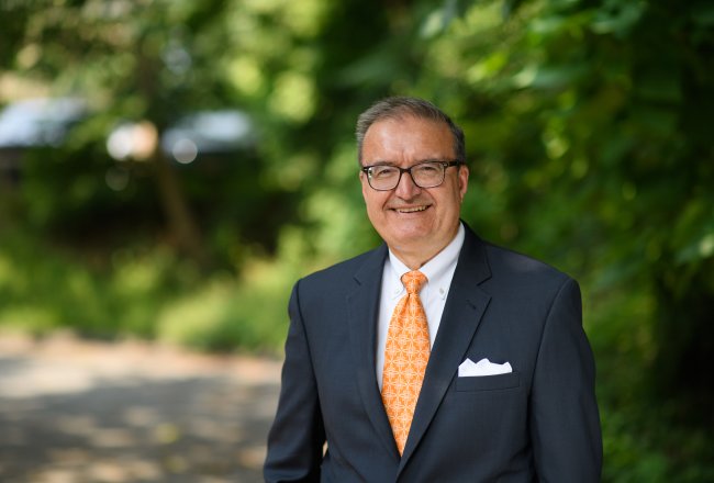 Tim McGowan in orange tie and blue blazer, stands outside with trees behind him and smiles at the camera.