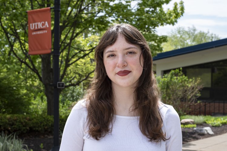 Bella Stephens, in white top with brown hair down past shoulders, stands in front of foliage on campus.